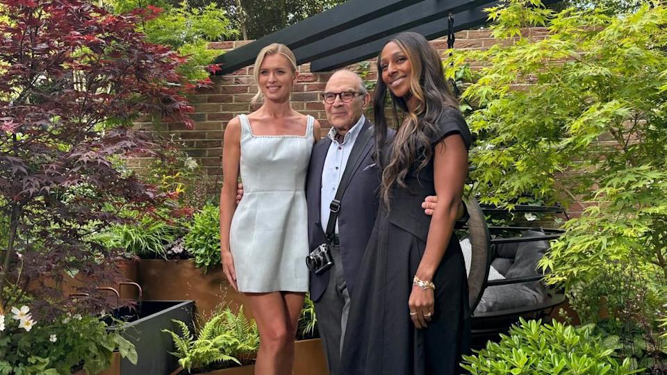 Two women in dresses are stood in the garden, an older man wearing a shirt and blazer is stood between the with his arms around theirs waists
