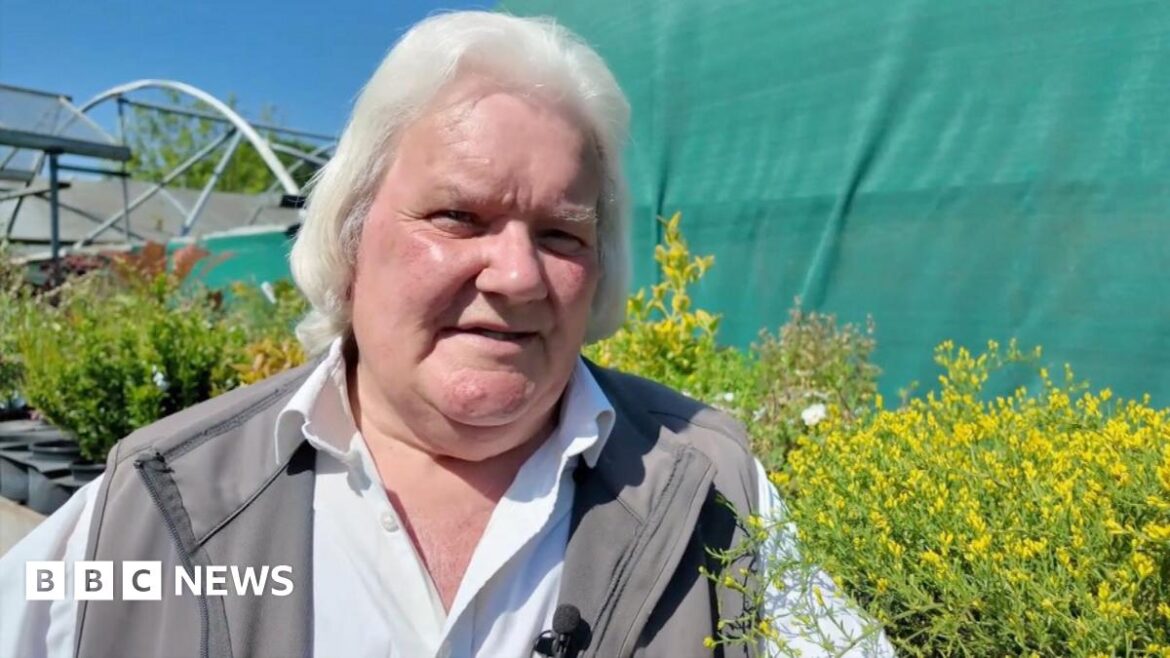 Stephen Goodridge is looking slightly off camera while giving a TV interview. He has mid length white hair and wears a white collared shirt with a grey waterproof unzipped gilet over the top. He stands in front of a row of evergreen shrubs and a green sheet in his garden centre.