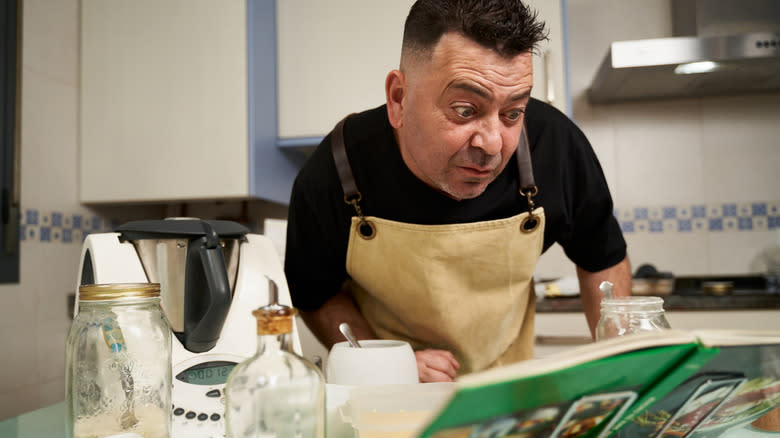 Man reading from a green covered cookbook propped up in a kitchen