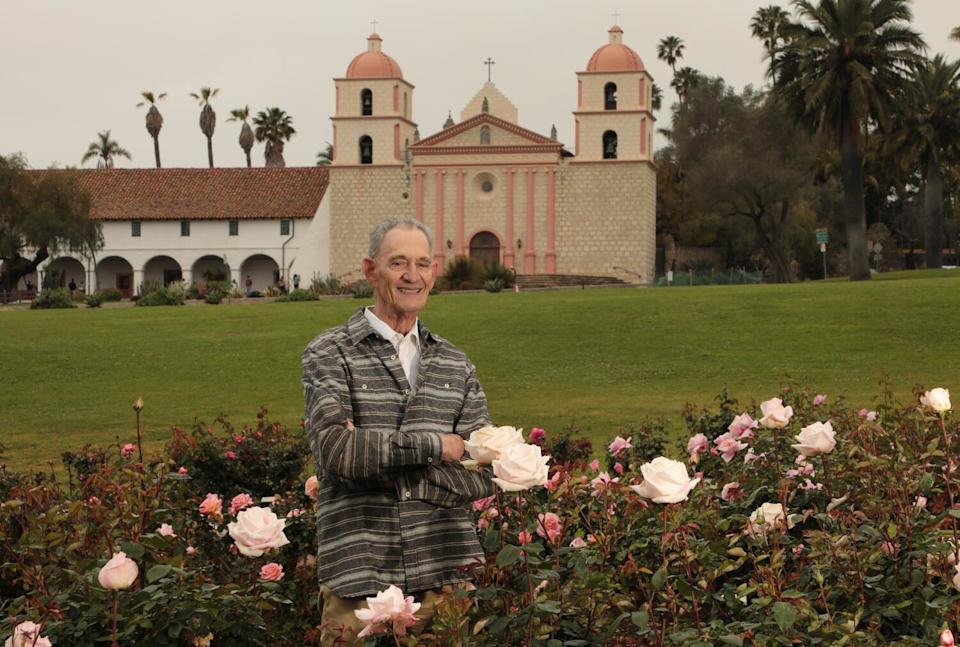 A tall slender man, Dan Bifano, standing in a bed of pale pink roses with a large pink-trimmed church in the background