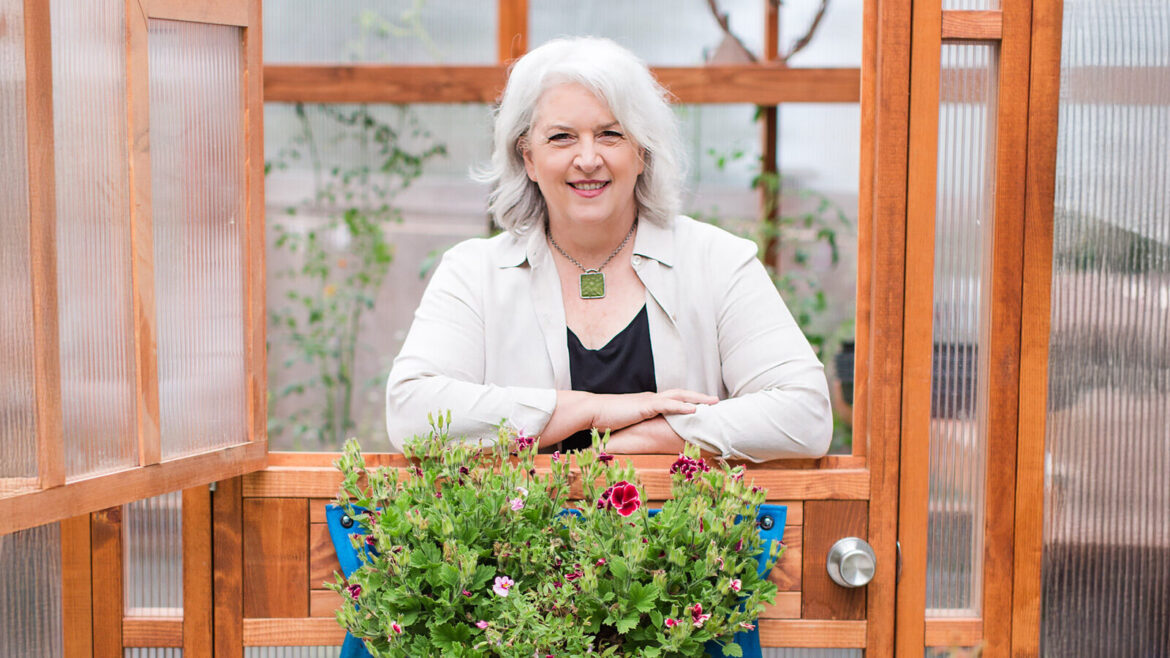 Not Flown, Grown: The Slow Flowers Movement is Having A Moment An older woman with gray hair stands smiling behind a wooden door, with blooming local flowers in front of her inside a greenhouse—a scene reminiscent of Debra Prinzing's focus on seasonal and sustainably harvested flowers.