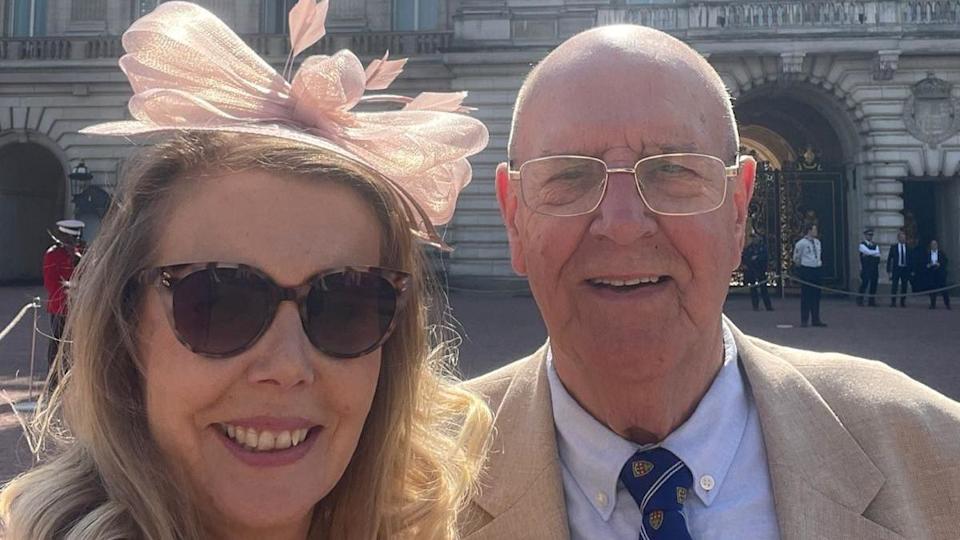 A male and female are taking a selfie - they are both smiling directly at the camera. The woman is blonde, wearing large tortoiseshell sunglasses and a pink fascinator. The male is bald, wearing thin silver glasses and a beige suit jacket with a blue shirt and navy tie.