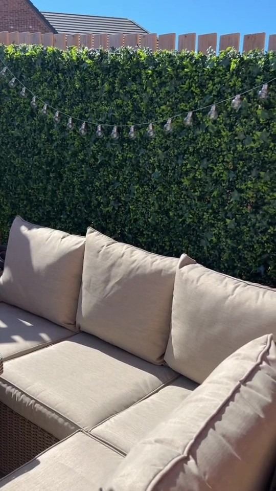 Beige patio sectional in front of artificial green wall with string lights.