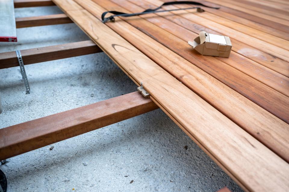 Close-up view of wooden deck boards being installed using hidden fasteners on a subframe. The image shows a construction phase with tools and material