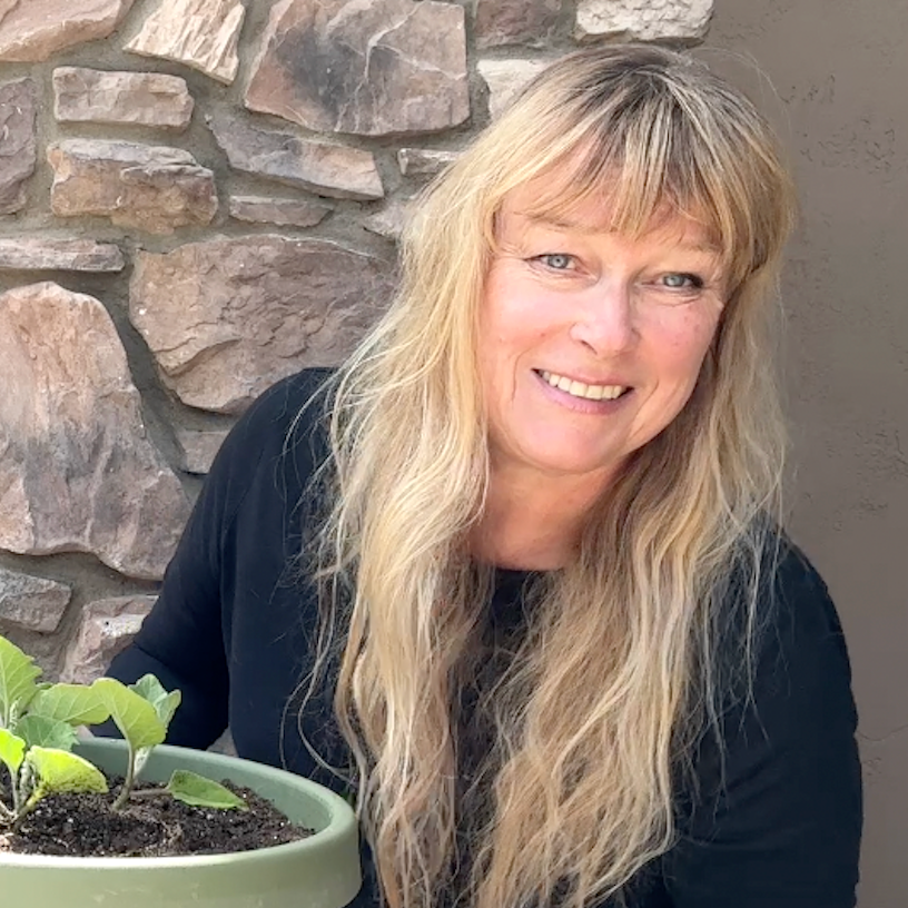 a light haired lady with a full fringe in a long sleeved black shirt kneeling down outside holding a green plant pot with a small plant inside