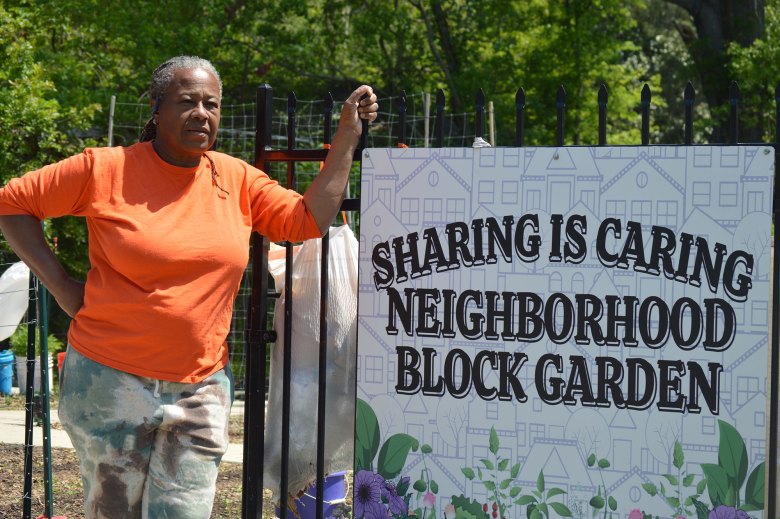 A woman in a bright orange shirt leaned next to a sign that reads "Sharing is Caring - Neighborhood Block Garden"