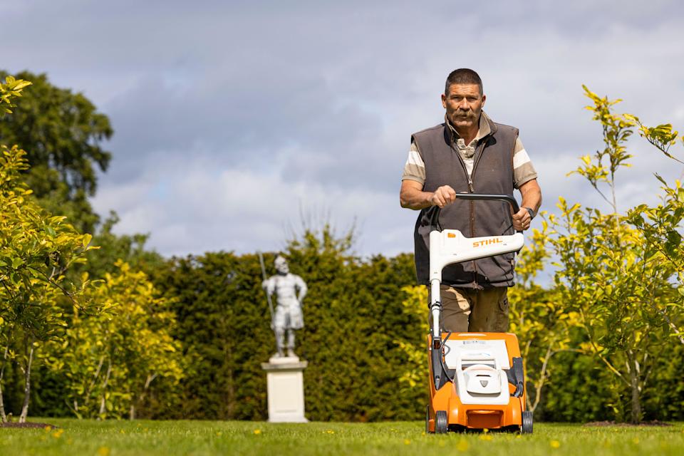 A man with a moustache and cropped dark hair pushes a lawnmower through a garden