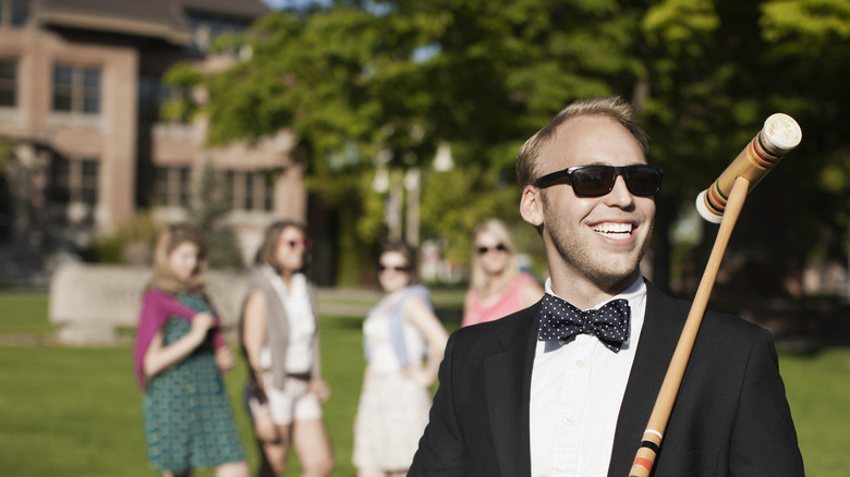 A man wearing a bowtie and sunglasses smiles while holding a croquet mallet