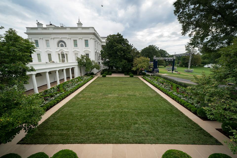 White House garden with trimmed lawn, flanked by trees and bushes, with a view of the building and a cloudy sky