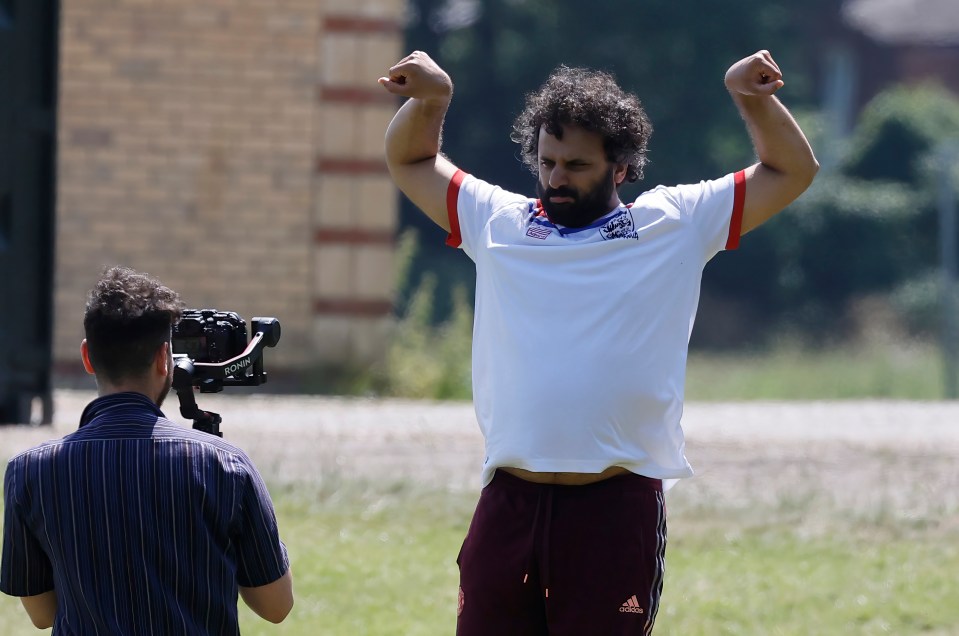 A man on a film set flexing his biceps while a cameraman films him.