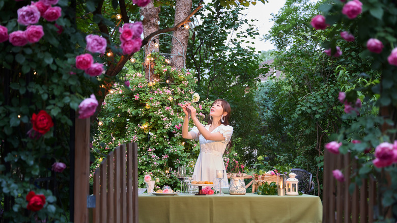 An elegantly dressed woman stands near a laden table surrounded by blooming roses at dusk