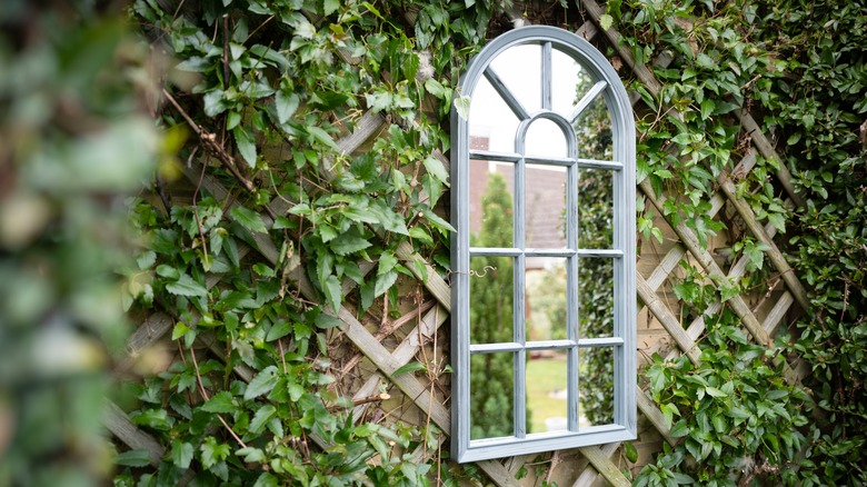 An arched mirror is hanging on a trellised wall in a garden
