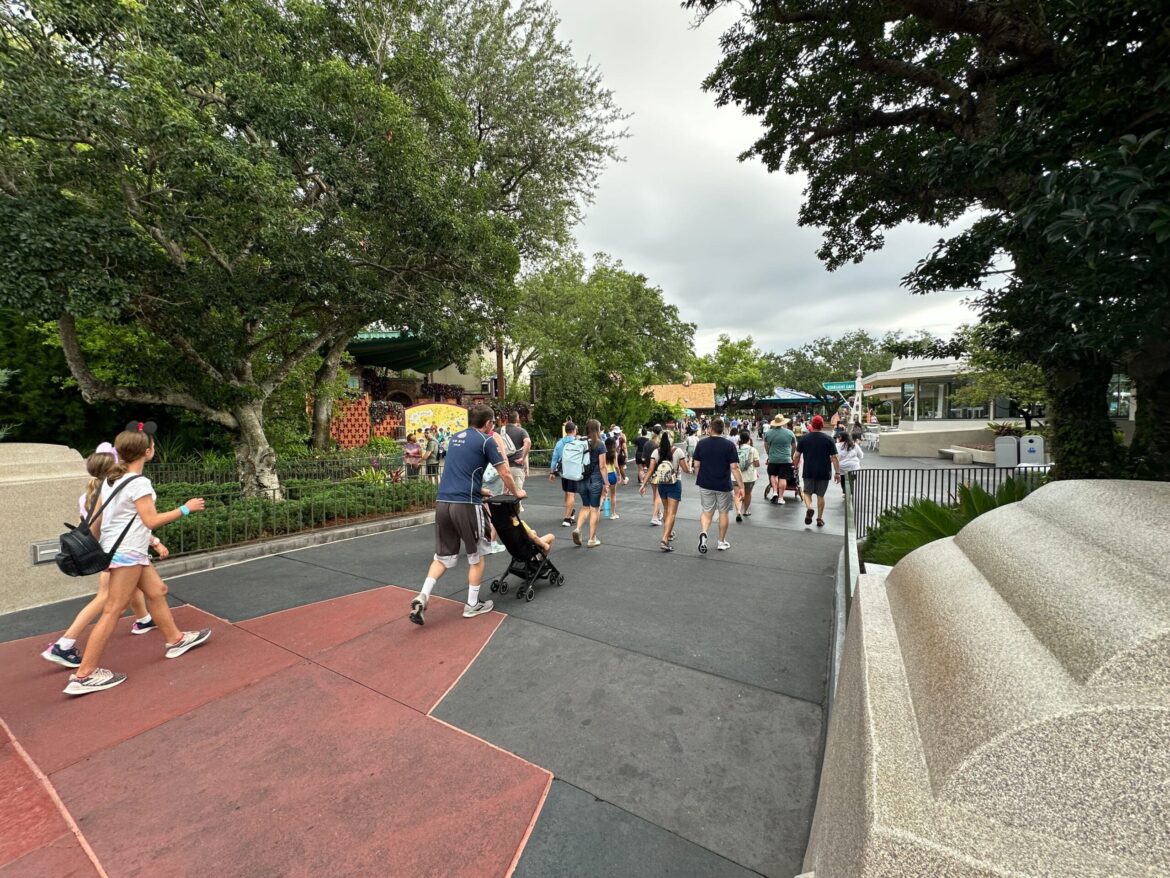 Pavement Replacement Seemingly Complete Between Cosmic Ray’s and Fairytale Garden in Magic Kingdom People walk along a wide pathway in a park with trees and greenery on a cloudy day.
