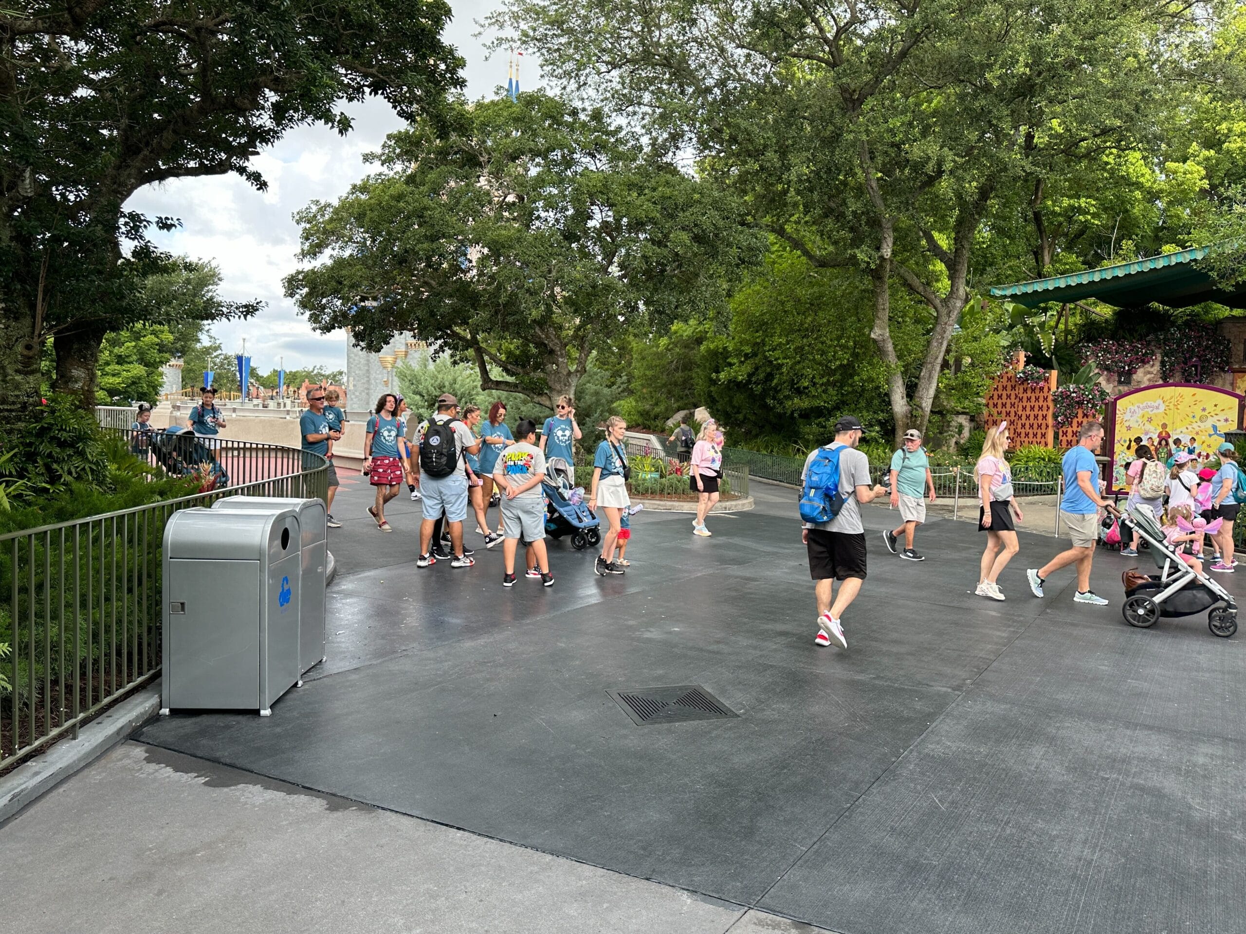 A group of people walk along a paved path lined with trees at a theme park on a cloudy day.