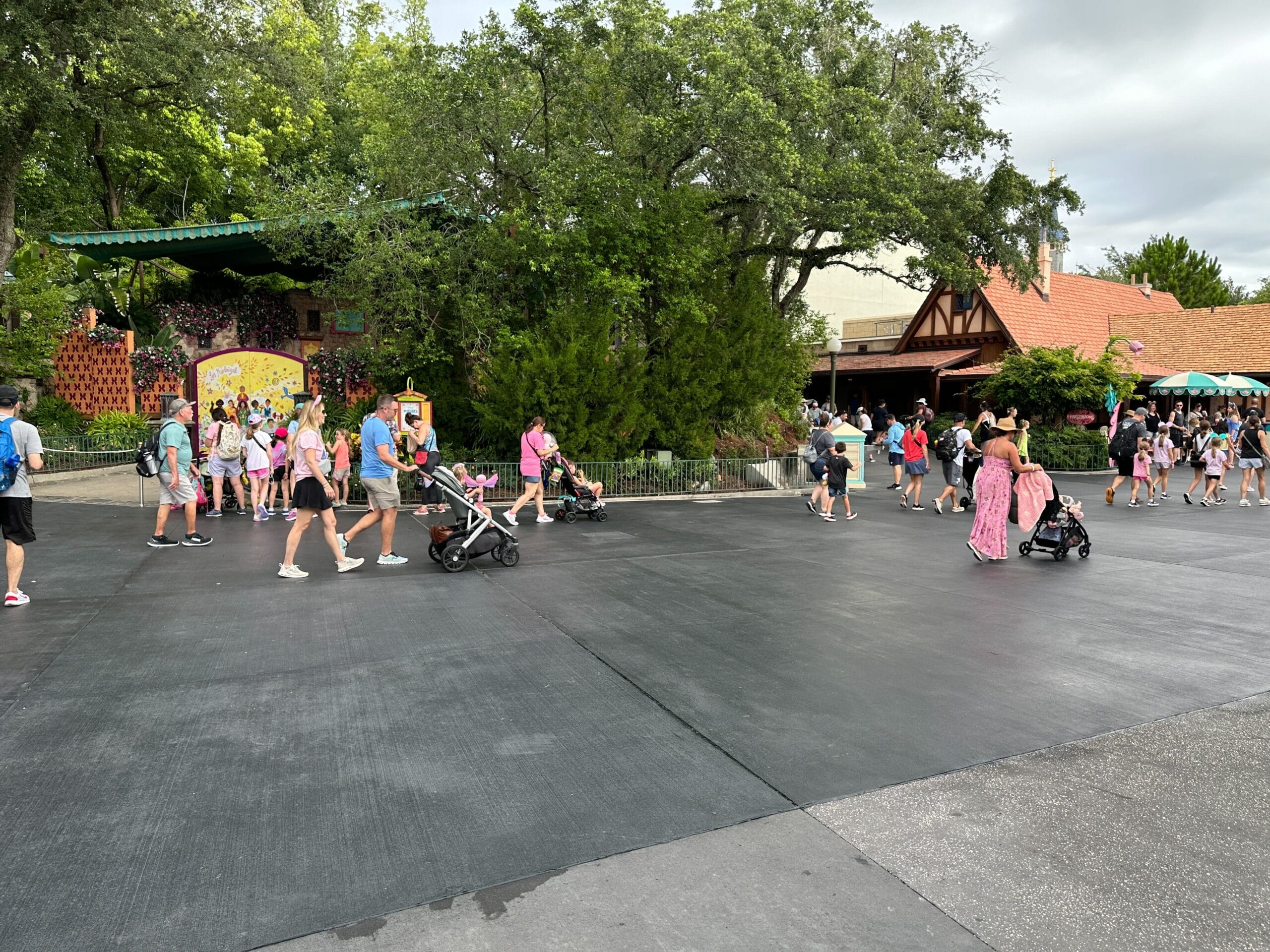 People walk with strollers near trees and buildings at a theme park on a cloudy day.