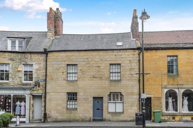 A stone cottage building with a dark blue front door in the centre set between shops in a terraced street.