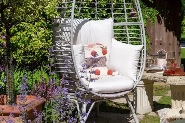 The chair with a rattan grey egg-shaped frame and white cushions inside, stood in a garden on a lawn surrounded by plants.