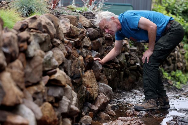 The image shows Jim, with white hair and wearing a blue t shirt, inside the deep brook and tending to the rocks in his stone wall