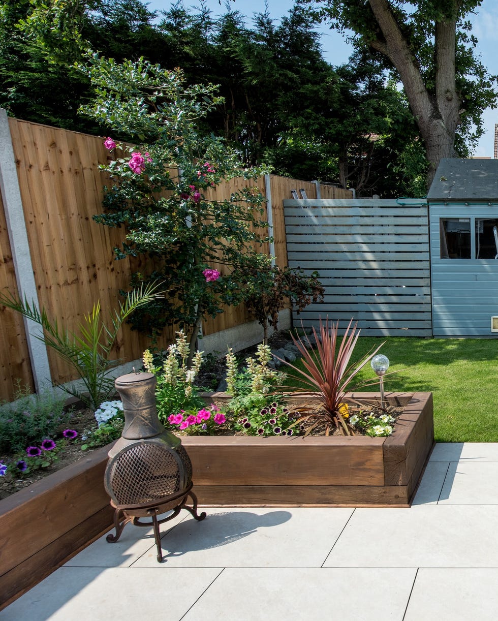 a general view of a back garden patio with white porcelain slabs, tiles, paving and wooden railway sleeper flowers beds leading on to a small grass lawn leading to a shed painted grey on a bright sunny day in summer