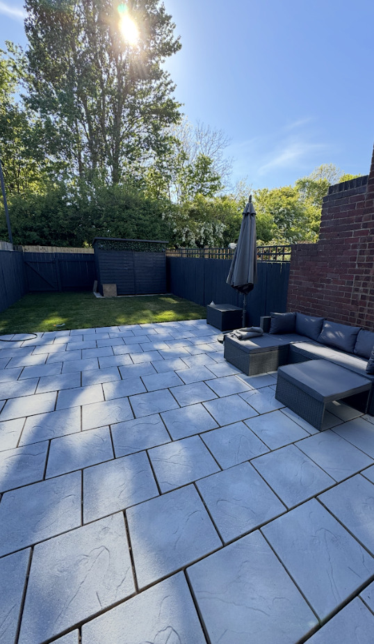 A renovated garden patio with light grey paving stones, a dark grey patio set, and a dark shed.
