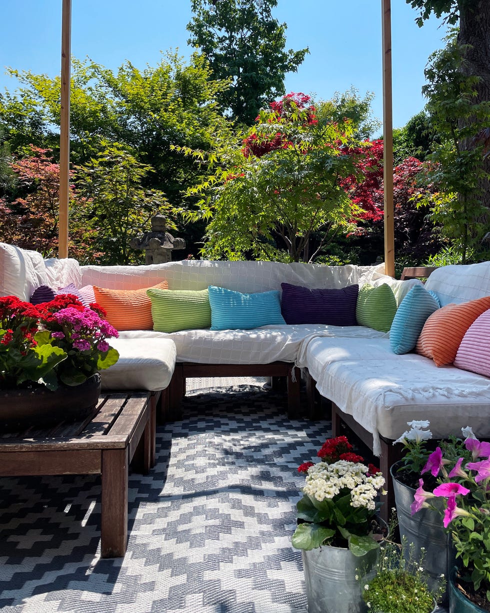 stock photo showing ornamental japanese style garden with outdoor lounge area on sunny day with fairy lights stung up on wooden pergola framework. featuring crystal clear koi pond, white tiled decking patio, japanese bonsai maples and hardwood, cushion covered seating.