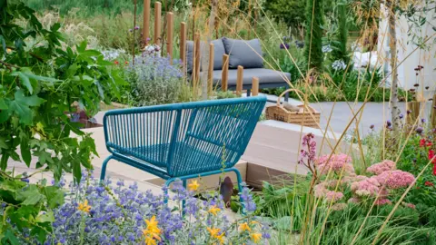 BBC Gardeners' World events/Jason Ingram A blue wicker chair is to the left of the image on light brown wooden decking. In the background is a grey seat which has two cushions. There are different coloured plants planted around the seating areas. 