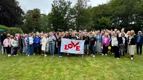 BBC News About 100 people holding a Coldplay love flag in the gardens at Holderness House. They're all smiling and grouped together.