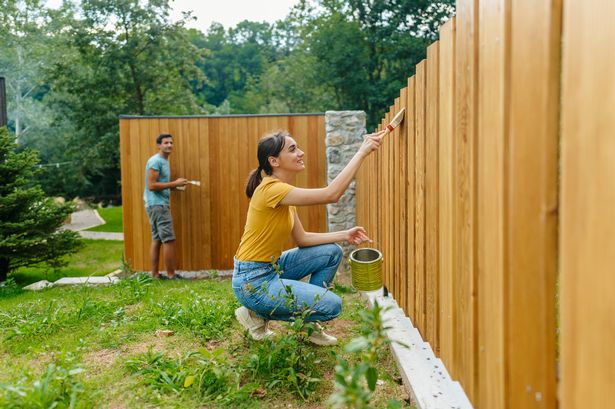 Photo of a young couple painting a picket fence and little wooden house in a yard.