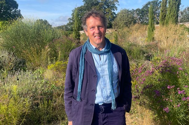 Still image from an episode of Gardeners World, January 2025, featuring presenter Monty Don. He is wearing a blue suit and light blue shirt, while standing in the middle of a field in the sunshine.