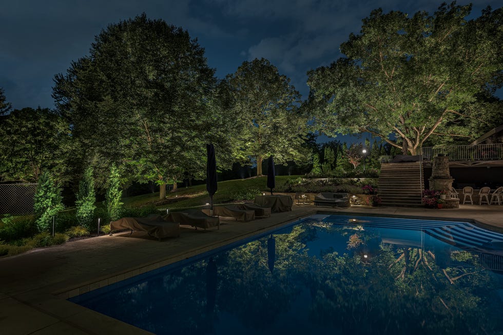Nighttime view of a well-lit outdoor swimming pool area surrounded by trees and landscape features.