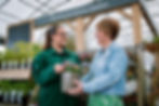 Woman holding silver plant pot with flowers reaching to young woman in garden centre 