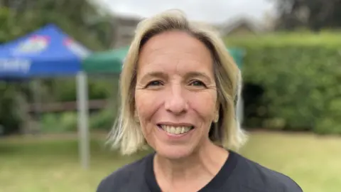 BBC News Woman with shoulder length blonde hair and a dark blue top. The background is blurred. She is standing on grass and there is a bush and a marquee behind her.