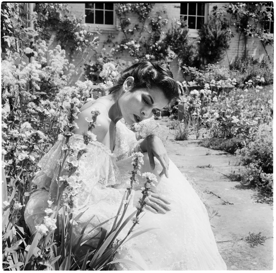 A black-and-white photograph of a woman in a lace gown sitting among tall flowers in a garden, leaning her head on her hand and looking down.