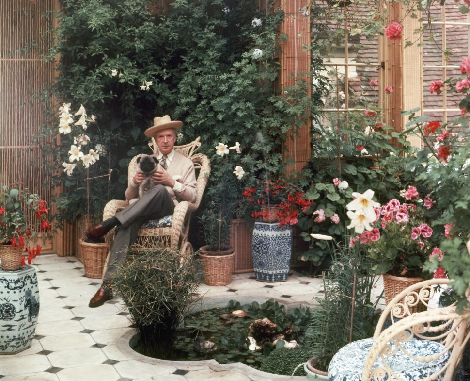 A color photograph of a man in a hat and suit sitting in a wicker chair holding a small dog in a plant-filled conservatory with potted flowers and a small pond.