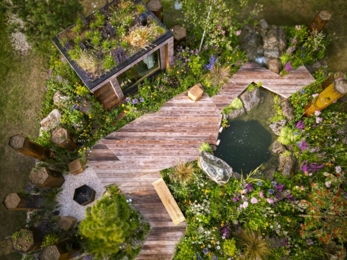 The Drakkar’s Drift garden. It has plants, flowers, bushes, grass and wooden structures and flooring. There is a pond to the right-hand side.