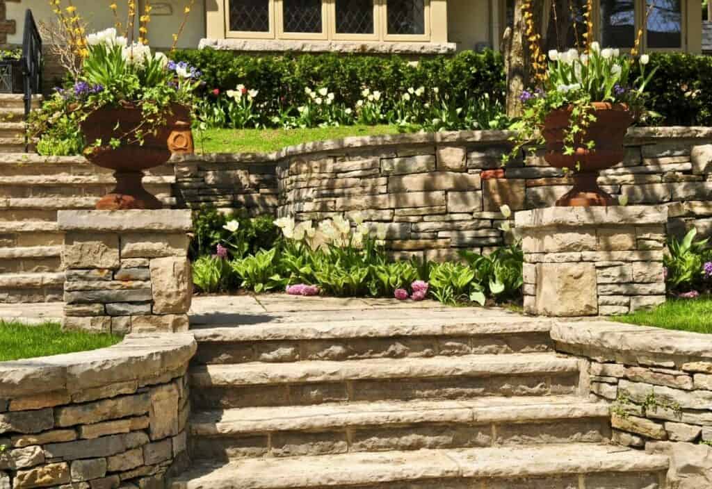Stone steps lead up to a tiered garden with fire safe landscaping, stone retaining walls, potted plants, and blooming flowers in front of a house.