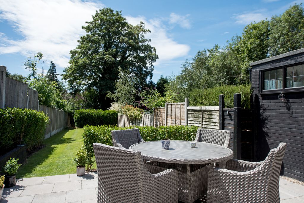 A general view looking down a back garden with a round grey rattan garden table and chairs on the patio on a sunny day with blue sky and white clouds