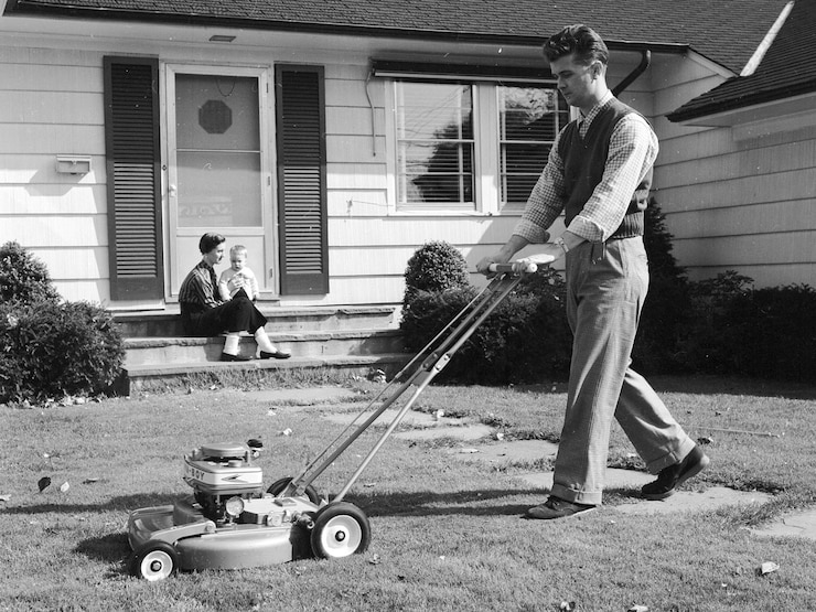 A black and white photograph of a man mowing the lawn as a woman and a toddler sit on the porch.