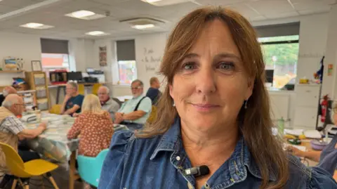 BBC Isla looks at the camera, she has a fringe and brown hair down to her shoulders and is wearing a denim top. Behind her a mix of people can be sat around a dining table, the majority of them are elderly.