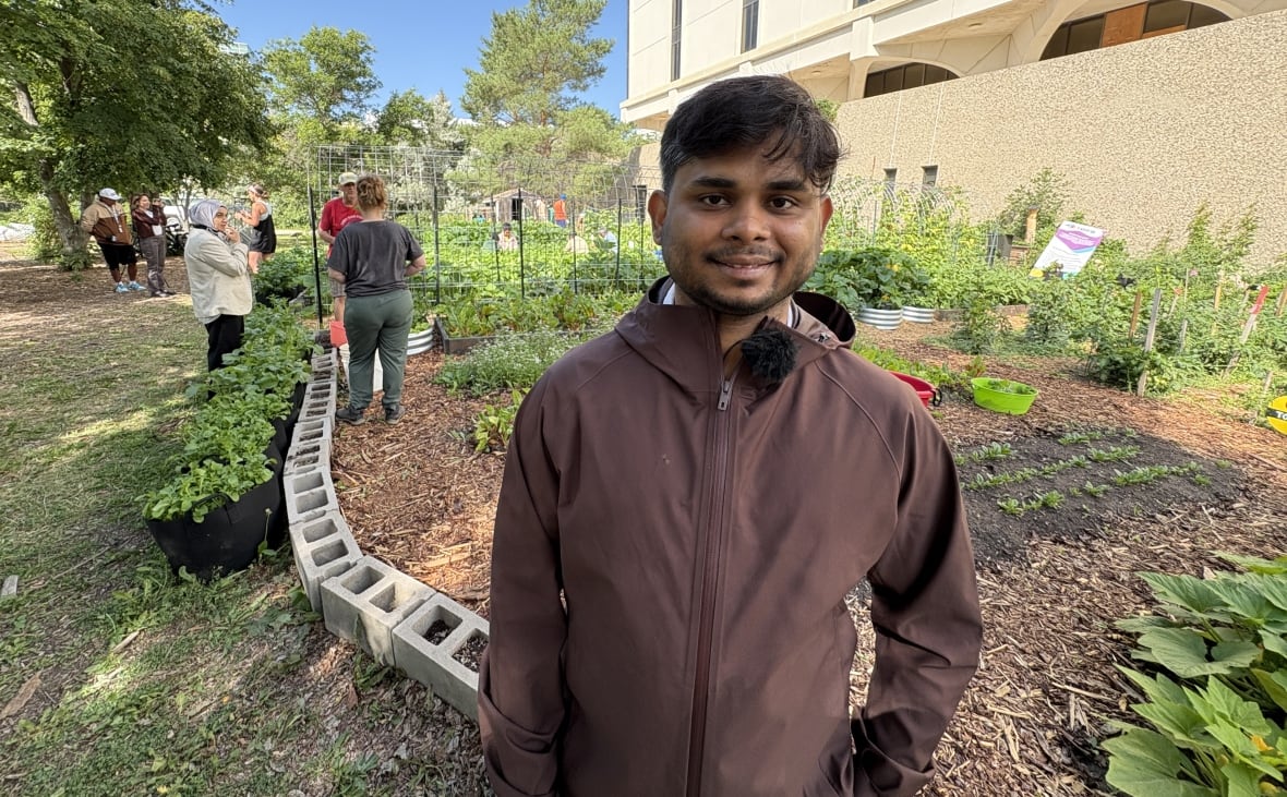 A smiling man in a brown jacket stands for a photo in a garden.