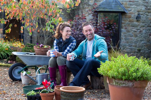 Couple taking a break from gardening. They smile while enjoying a drink with pots, plants and gardening tools. You can see the leaves starting to change colour in the background.