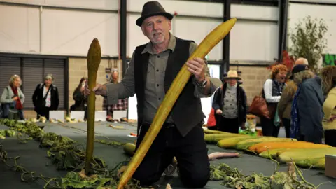 Submitted A man is kneeling on a table filled with display vegetables. He is holding two long fruits from a luffa he has grown, which are tube-like and yellow in colour. They appear to be about a metre long. The vine of the plant is laid out on the table behind him, infront of him and around him.