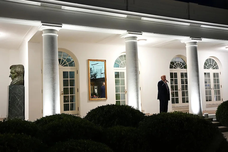 A person stands outside a brightly lit building at night, near large columns and shrubbery, next to a statue bust and framed portrait