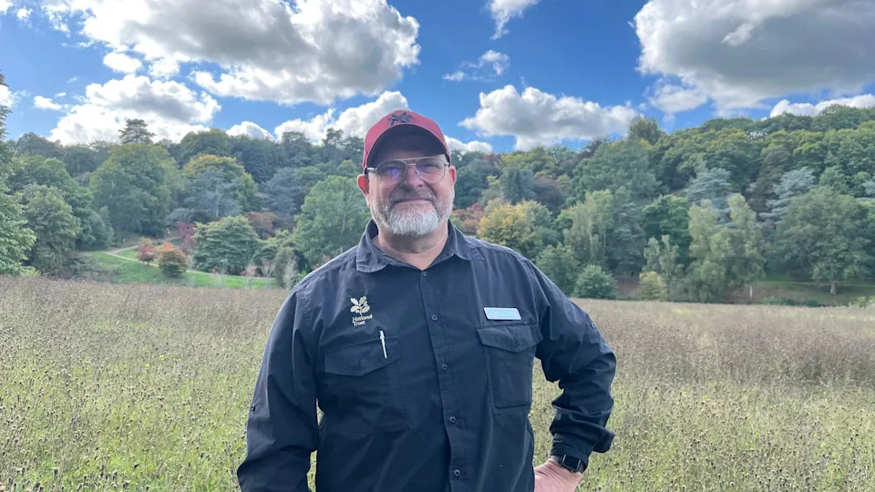 A man in a dark shirt with the National trust logo on it and a red cap. He is stood in a grassy meadow with a series of trees in the background, some of which are turning red.