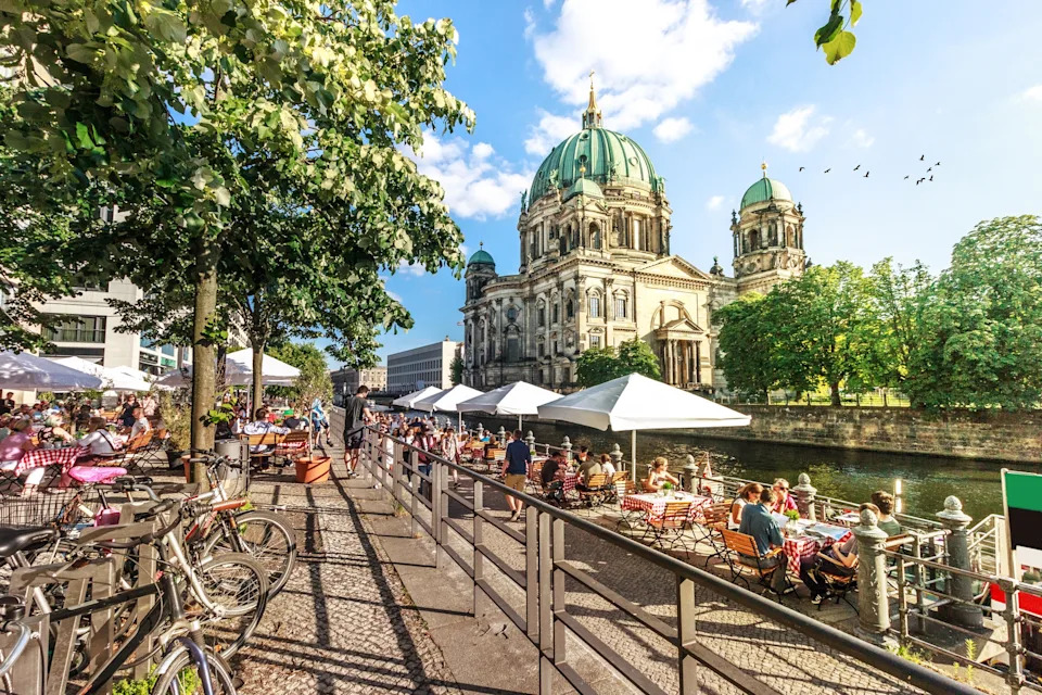 People dining outdoors by a canal with bicycles and trees nearby, and a large cathedral in the background