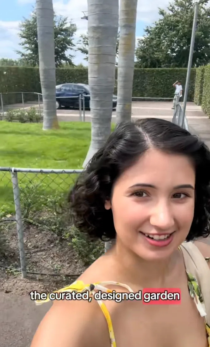 Person taking a selfie in a garden with palm trees and lush greenery. Text at the bottom reads: "the curated, designed garden."