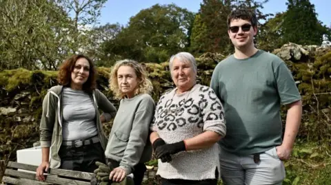 BBC Four people, the volunteers, standing in a row. From left to right: a woman with long dark curly hair, wearing a green fleece and a grey t-shirt; a woman with curly fair hair, wearing a light green sweatshirt; a woman with short grey hair, wearing a light coloured sweater with sleeves rolled up, and dark gloves; a young man with short dark hair, wearing sunglasses and a short-sleeved green t-shirt.