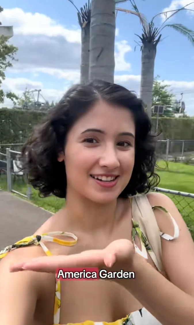 Person outdoors smiling for a selfie, gesturing towards a park area with palm trees. Text on image reads "America Garden."