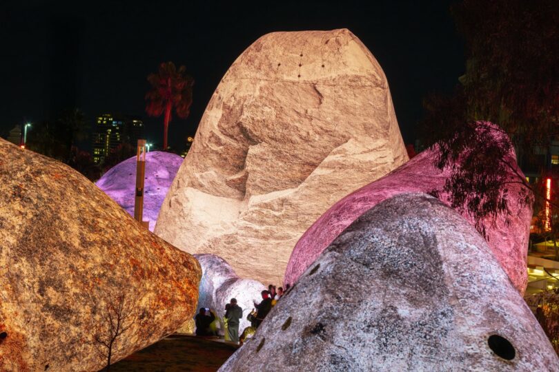Several large boulders illuminated in pink and white light, with people standing and walking around them in an urban outdoor setting at dusk