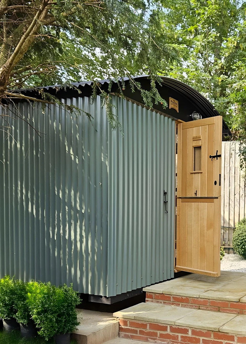 a green utility hut with a wooden door, brick steps next to it, and a leafy green bush